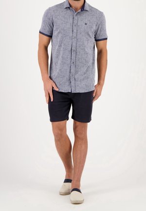 Mand iført lyseblå kortærmet skjorte med knapper, mørkeblå shorts og beige slip-on sko, der går på hvid baggrund.