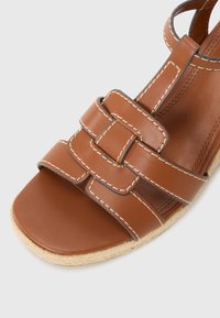 Brown leather sandal featuring woven straps, contrast stitching, open toe, and jute-wrapped sole. Smooth texture with rounded edges.