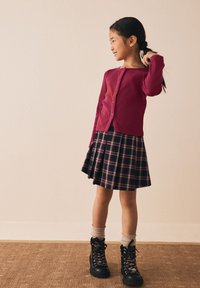 Bordeauxrode cardigan met knopen gecombineerd met een geblokte skaterrok in donkere tinten. Zwarte veterlaarzen met grijze sokken. Glad interieur als achtergrond.