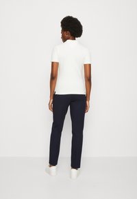 Camicia bianca a maniche corte con scollatura tonda, abbinata a pantaloni blu navy e sneakers bianche. La camicia presenta aperture laterali per facilitare i movimenti.