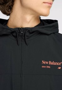 Schwarze New Balance Jacke mit durchgehendem Reißverschluss, verstellbarem Kapuzenkragen und korallenfarbener Logo-Aktion; glatte Textur mit nahtloser Verarbeitung.