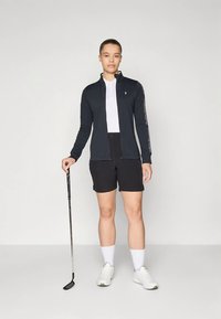 Golfbeklædning med en sort lynlåstrøje med hvide detaljer, en hvid polo og sorte shorts. Modellen holder en golfkølle.