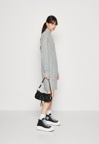Grå strikket kjole med høy hals og lange ermer, parret med svarte høyhælte sneakers og en svart håndveske med sølvacenter.