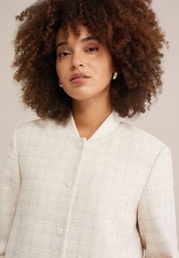 Veste claire texturée avec un col lisse, comportant des boutons ronds et un détail de poche simple.