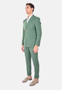 Abito doppiopetto in lino verde con bottoni dorati, pantaloni slim fit e maniche arrotolate, abbinato a mocassini grigio chiaro. Camicia bianca sotto.