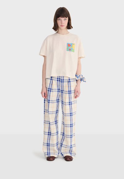 Femme portant un t-shirt court crème avec un motif floral coloré, un pantalon crème à jambes larges avec un motif à carreaux bleu et orange, et des sandales marron ouvertes à l'avant.