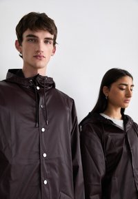 Brune, vanntette jakker med hette, med front trykknapper, snøring og sidelommer. Stoffet har en jevn, blank tekstur.