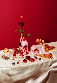 Różowe klapki w serca, butelka szampana, róża, kawałki pizzy i różne jedzenie na białym obrusie.