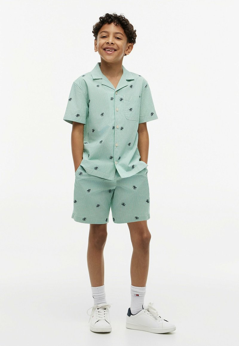Garçon souriant, portant une chemise à manches courtes boutonnée vert clair et un short assorti avec des motifs d'abeilles noires, des chaussettes blanches et des baskets blanches.
