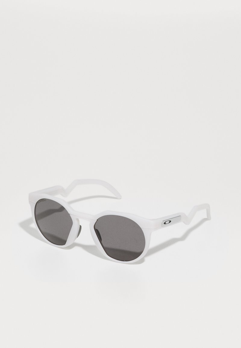 Gafas de sol de plástico blanco con lentes octagonales, lentes gris oscuro y un diseño único de patillas en zigzag; presenta un pequeño logotipo plateado en el costado.