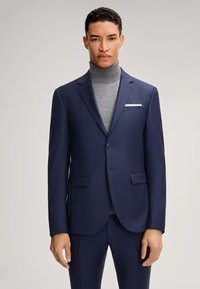 Marineblauer Anzug mit schmaler Passform, strukturierter Oberfläche. Mit Revers, zwei aufgesetzten Taschen vorne, einem einknopfigen Verschluss und einem weißen Taschentuch als Akzent.