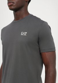 Grå bomulls t-skjorte med rund halslinning og korte ermer. Har en hvit EA7 Emporio Armani-logo på venstre brystområde.