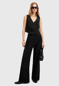 Schwarze ärmellose Weste mit fünf Knöpfen, kombiniert mit weit geschnittenen schwarzen Hosen. Glattes Material mit minimalistischem Design; trägt eine schwarze Handtasche.