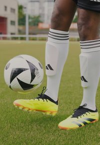 Botas de fútbol amarillas con acentos negros y una superficie texturizada, combinadas con calcetines blancos con rayas negras, se encuentran posicionadas cerca de un balón de fútbol en blanco y negro.