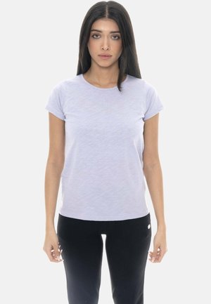 Donna con lunghi capelli scuri che indossa una maglietta viola chiaro a maniche corte e pantaloni neri, in piedi su sfondo bianco.