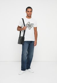Hvid T-shirt med sort og beige grafik; blå jeans; sort skuldertaske; hvide sneakers. Model står mod en ensfarvet baggrund.