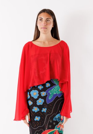 Donna con lunghi capelli castani indossa una maglia ampia rosso brillante a maniche lunghe e una gonna a vita alta con fantasia floreale.