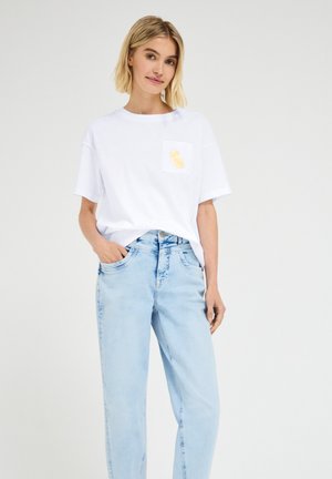 Kvinde iført en hvid t-shirt med en citron grafik på lommen og lyseblå højtaljede jeans, stående med den ene hånd i lommen.