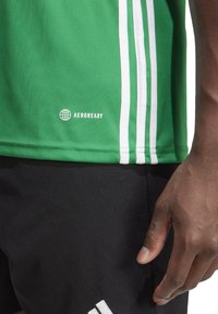 Maglietta sportiva verde con strisce bianche, realizzata in tessuto traspirante. Presenta il logo "AEROREADY" e un design aderente. Pantaloni corti neri visibili sotto.