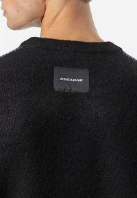 Suéter preto de malha texturizada com decote redondo. Apresenta uma etiqueta cosida na parte de trás com a palavra "PEGADOR" em letras brancas.