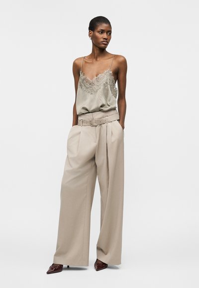 Γυναίκα που φοράει μπεζ καλοκαιρινή μπλούζα με δαντέλα, παντελόνι ψηλόμεσο, φαρδύ με ζώνη και καφέ μυτερά τακούνια, στέκεται με τα χέρια στις τσέπες.