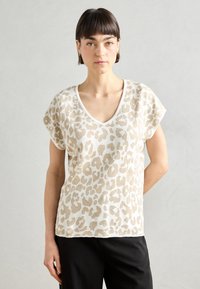 Haut en imprimé léopard beige avec un col en V et des manches courtes, fabriqué à partir d'un tissu doux ; associé à un pantalon noir, sur un fond neutre.