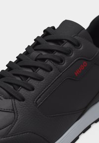 Μαύρο δερμάτινο αθλητικό παπούτσι με μαύρα κορδόνια, υφασμένα πλαϊνά πάνελ, λευκή σόλα και κόκκινο λογότυπο «HUGO» στο πλευρό.