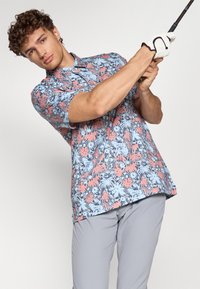 Short-sleeve floral golf shirt with a blue background, featuring pink and black patterns. Worn with gray golf pants and a white glove.