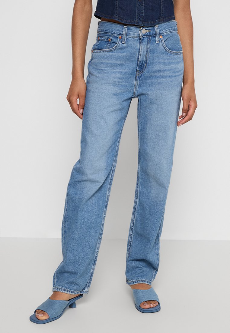Hellblaue Denim-Jeans mit geradem Schnitt und klassischem Fünf-Taschen-Design. Mit einem Reißverschluss und Gürtelschlaufen ausgestattet, kombiniert mit blauen, offenen Mules.