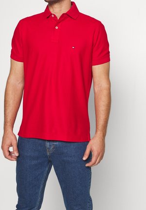 Mężczyzna w jaskrawoczerwonej koszulce polo z krótkim rękawem i małym logo na piersi oraz niebieskich jeansach, stojący na jednolitym białym tle.