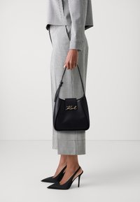 Sac à main en cuir noir avec logo doré, forme structurée et fine bandoulière. Modèle portant un costume à carreaux et des escarpins noirs.