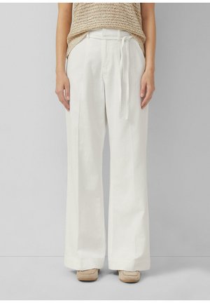 Person trägt weiße, weit geschnittene Hose mit hohem Bund und Stoffgürtel, beiges Stricktop und beigen Schuhen, steht vor einem schlichten grauen Hintergrund.