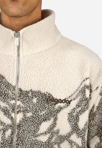 Kurtka polarowa z wysokim kołnierzem w kolorze kremowym i szarym, z zamkiem błyskawicznym i teksturowanymi wzorami. Logo umieszczone po lewej stronie.