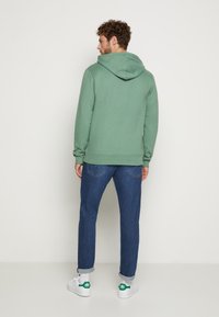 Grünem Kapuzenpullover mit gerippten Bündchen, kombiniert mit dunkelblauen Jeans und weißen Sneakers mit grünen Akzenten, von hinten betrachtet.