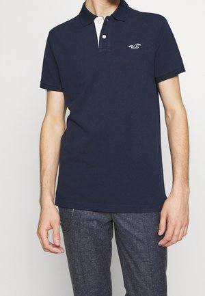 Homme portant un polo bleu foncé à manches courtes avec un petit logo blanc sur la poitrine et un pantalon gris avec un accessoire chaîne en métal.