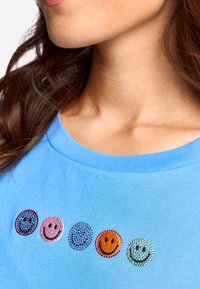 Blå bomullströja med fem färgglada strassleende ansikten i rad, med ribbad rund halsringning och slät textur.