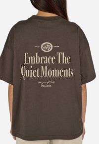 Brązowy oversize t-shirt z krótkim rękawem. Zawiera duży biały napis na plecach: "Przyjmij Ciche Chwile" oraz szczegóły logo.