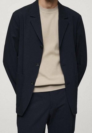 Personne portant un costume bleu marine foncé avec un blazer boutonné par-dessus un pull en maille beige, debout devant un fond uni clair.