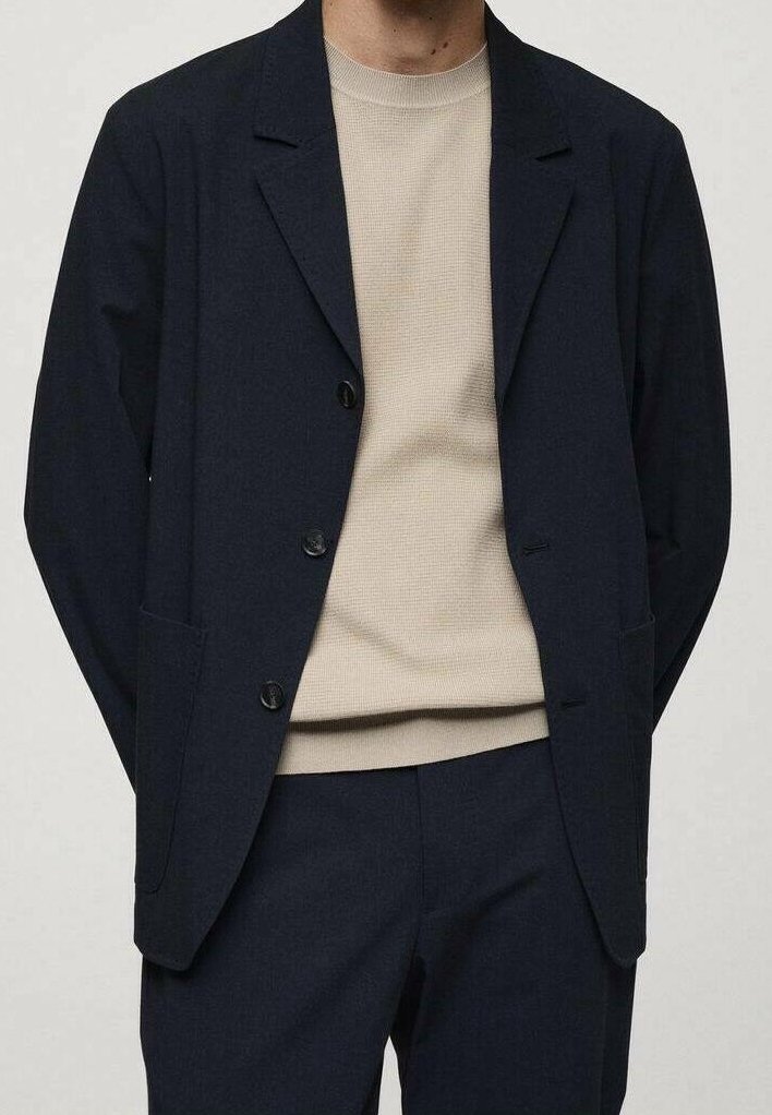 Personne portant un costume bleu marine foncé avec un blazer boutonné par-dessus un pull en maille beige, debout devant un fond uni clair.