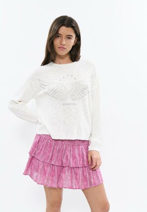 Mujer con cabello largo y oscuro que lleva un suéter blanco con diseño de alas y una falda rosa de capas, de pie contra un fondo blanco.