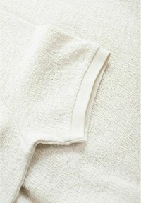 Weißer strukturierter Stoff mit einem subtilen Muster, ausgestattet mit einem weichen Strickbund. Nahaufnahme des Ärmeldetails am Hauptkleidungsstück.