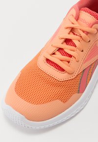 Scarpa da ginnastica arancione con tomaia in rete, sovrastrutture testurizzate e accenti rossi a contrasto. Presenta una suola bianca e chiusura con lacci.