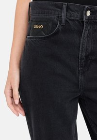 Jeans in denim nero con una tasca trapuntata nera con il logo "LIU-JO" in oro. Il tessuto ha una texture liscia e un design slim fit.