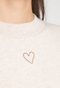 Svetle béžová mikina vyrobená z měkké látky s kulatým výstřihem. Na hrudi má malé hnědé vyšívané srdíčko.