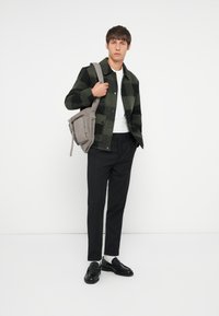 Grünes und schwarzes karriertes Flanelljackett, weißes Hemd, schwarze Hose, schwarze Mokassins und eine graue Schultertasche mit Reißverschluss-Detail.