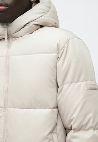 Béžová prešívaná bunda s kapucňou, s prešívanými časťami, predným zipsom a značkovou nálepkou na ľavom rukáve. Hladká textúra.