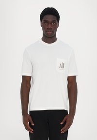 Biela bavlnená tričko s krátkymi rukávmi, okrúhlym výstrihom a prednou vreckom s hnedým grafickým logom. Model stojí na neutrálnom pozadí.