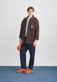 Struktureret brun overskjorte, lyseblå sweater, navy bukser, orange sko og kasket. Carhartt-logo på skjortens lomme. Model står foran en blå baggrund.