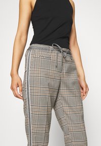 Lehké kostkované kalhoty v béžové a černé barvě, se stahovací šňůrkou v pase, bočními kapsami a bílými kontrastními pruhy po stranách.