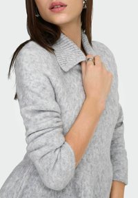 Šedý pletený svetr s žebrovaným límcem a 3/4 rukávy, s jemnou texturou a nenápadným vzorem. Ruka zdobená stříbrným prstenem.